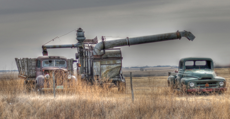 A machine and two pickup trucks from the 1940s or 1950s sit in a field